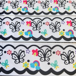 May include: White ribbon with a black scalloped edge. The ribbon has a repeating pattern of black butterflies, rainbows, stars, and flowers. The colors in the pattern are pink, yellow, blue, green, and purple.