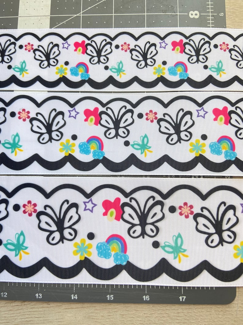 May include: White ribbon with a black scalloped edge. The ribbon has a repeating pattern of black butterflies, rainbows, stars, and flowers. The colors in the pattern are pink, yellow, blue, green, and purple.