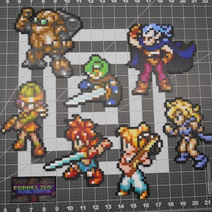 Puede incluir: Un conjunto de siete personajes de pixel art, cada uno hecho de cuentas de colores. Los personajes son un robot, un guerrero, un mago, un caballero, un ladrón, un sanador y un dragón. Los personajes están todos en diferentes poses y tienen diferentes armas y accesorios.