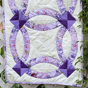 以下が含まれることがあります： 蝶と花柄のダブルリングデザインが施された、パープルとホワイトのキルトの毛布。このキルトはさまざまな生地で作られており、パープルの縁取りで仕上げられています。