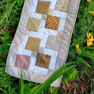 May include: A beige table runner with a patchwork design featuring various shades of brown and beige fabrics. The runner is made of squares with different patterns and textures.