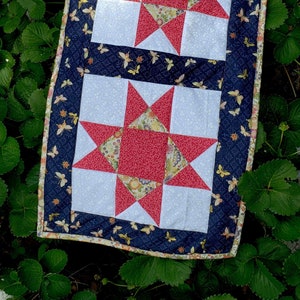 Może przedstawiać: Ręcznie robiony koc z granatową lamówką i białym tłem. Koc zawiera dwa bloki gwiazd w kolorze czerwonym i białym z kwiatowym wzorem. Koc idealnie nadaje się do sypialni lub salonu.