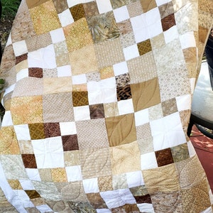 May include: A quilt made of various shades of brown and beige fabric squares with a white background. The quilt is quilted with a leaf pattern.