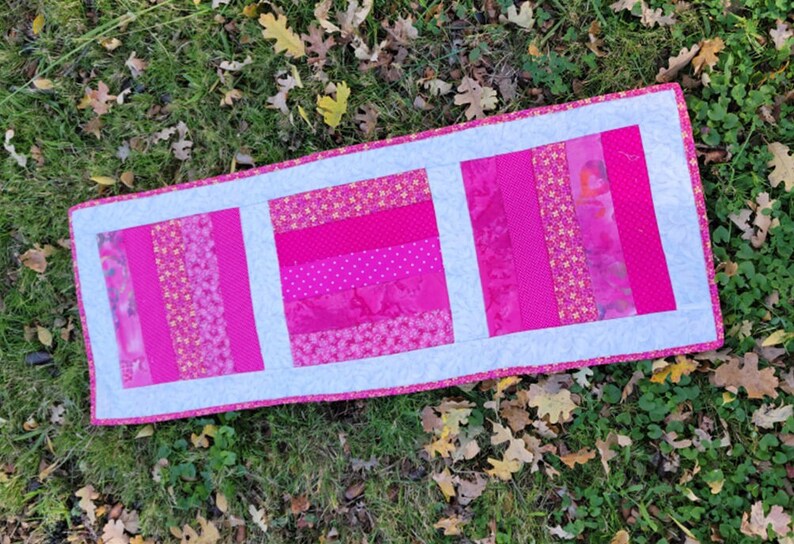 Pu&ograve; includere: Un runner da tavolo rosa e bianco con un design a patchwork. Il runner &egrave; realizzato con diversi tessuti con motivi floreali e geometrici.