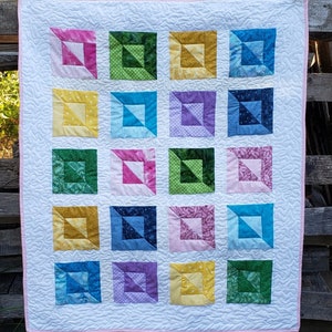 May include: A white quilt with a pink border. The quilt is made up of 16 squares, each with a different patterned fabric. The patterns include polka dots, floral prints, and geometric designs. The colors are bright and cheerful, including pink, yellow, green, blue, and purple.