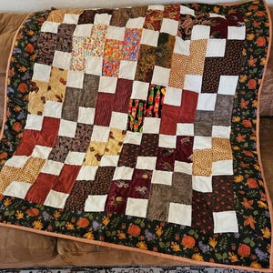 May include: A patchwork quilt with a checkerboard pattern. The quilt features squares of various fabrics in shades of brown, red, yellow, and white. The border is a dark green with a floral and pumpkin design. The quilt is displayed on a brown couch.