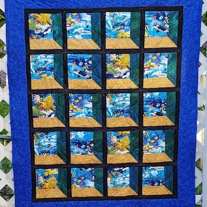 Może przedstawiać: Niebieska kołdra z efektem 3D, przedstawiająca 16 kwadratów z różnymi scenami podwodnymi. Każdy kwadrat ma czarną i złotą ramkę. Sceny przedstawiają delfiny, rafy koralowe i ryby.