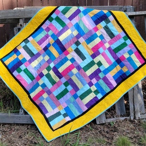 May include: A colorful quilt with a patchwork design featuring squares of various colors, including pink, blue, green, yellow, and purple. The quilt is bordered with a wide yellow band and a black trim.