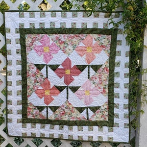 May include: A white quilt with a green border and a floral pattern. The quilt features pink and white flowers in a geometric design.