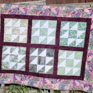 Könnte beinhalten: Eine handgefertigte Steppdecke mit floralem Rand und sechs quadratischen Blöcken. Jeder Block zeigt ein Sternmuster in Grün-, Weiß- und Grautönen. Die Steppdecke ist von einem dunkelburgunderfarbenen Rand umgeben.