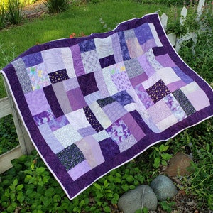 May include: A purple patchwork quilt with a variety of shades of purple and white fabric squares. The quilt is made with a variety of different patterns and textures.