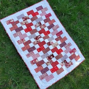 May include: A white quilt with a red and white checkered pattern. The quilt is made up of squares of different fabrics, each with a different floral print. The quilt is lying on a bed of green grass.
