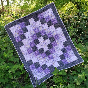 May include: A purple and white patchwork quilt with a checkered pattern. The quilt is made of squares of fabric with different floral designs. The quilt is hanging on a fence.