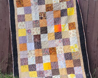 Può includere: Una trapunta patchwork con un motivo a griglia di quadrati in vari colori e motivi. La trapunta presenta quadrati nelle tonalità di giallo, arancione, marrone e viola. Il bordo è nero con un disegno a foglie e la trapunta è appoggiata a una staccionata di legno.