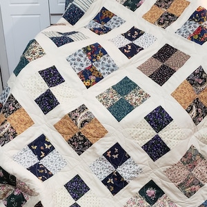 Peut inclure: Une courtepointe de couleur crème avec un motif de patchwork composé de carrés de différents motifs floraux et paisley dans des tons de bleu, vert, marron et beige. La courtepointe est bordée d'une bordure florale bleue et blanche.