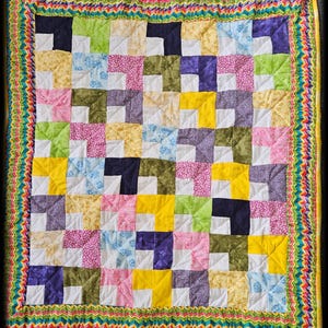 May include: A patchwork quilt in a variety of colours, including pink, yellow, blue, green, and purple. The quilt has a grid pattern of squares. The border is a zig-zag pattern in multiple colours. The quilt is likely made of cotton.