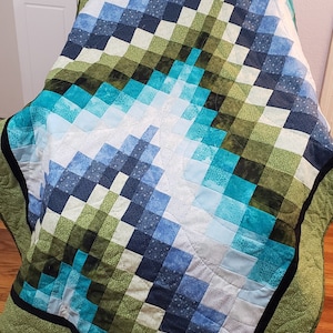 Puede incluir: Una colcha de retazos verde, azul y blanca con un borde negro. La colcha está doblada y sentada en un suelo de madera.