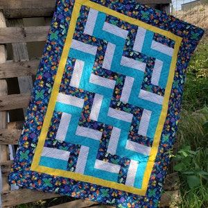 Op de afbeelding: Een blauwe en witte quilt met een gele rand en een patroon van vlinders en bloemen. De quilt is gemaakt van vierkante stoffen die in een geometrisch patroon aan elkaar zijn genaaid.
