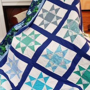May include: A blue and white patchwork quilt with a star pattern. The quilt is made of different fabrics and has a blue border. The quilt is folded and draped over a chair.