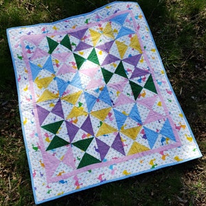 Op de afbeelding: Een kleurrijk quilt met een geometrisch patroon van driehoeken in tinten roze, blauw, groen, geel en paars. De quilt is gemaakt van katoenen stof en heeft een witte achtergrond met kleine bloemenmotieven.