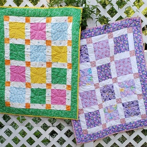 Pode incluir: Duas colchas de retalhos feitas à mão com uma variedade de tecidos coloridos. A colcha à esquerda tem uma borda verde e um fundo branco com quadrados amarelos, rosa e azuis. A colcha à direita tem uma borda roxa e um fundo branco com quadrados roxos e brancos.