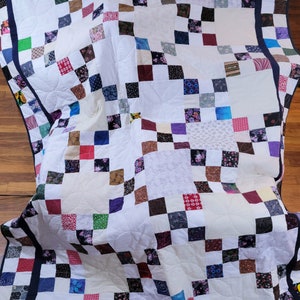 Könnte beinhalten: Eine weiße Steppdecke mit Patchwork-Muster, das aus einer Vielzahl von bunten Quadraten besteht. Die Steppdecke ist mit einem dunkelblauen Rand eingefasst und hat ein gestepptes Muster.