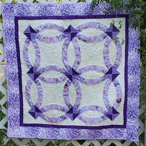 Op de afbeelding: Een paarse en witte quilt met een dubbele ringontwerp. De quilt is gemaakt van stof met een bloemenpatroon en is gequilt met een swirl-ontwerp.