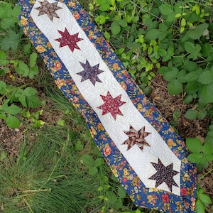 Może przedstawiać: Biegacz na stół w kolorze niebieskim i białym z kwiatowym wzorem i rzędem pięciu gwiazdkowych łatek z tkaniny w kolorze czerwonym, brązowym i niebieskim.