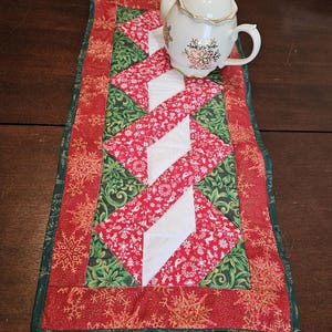 Puede incluir: Un camino de mesa navideño festivo con un diseño de patchwork rojo, verde y blanco. El camino de mesa presenta un patrón repetido de copos de nieve y un patrón central en zigzag. Una tetera blanca con detalles dorados encima.