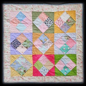 May include: A square patchwork quilt featuring a grid of nine large blocks, each containing a smaller diamond-shaped center made of four distinct fabric squares. The quilt displays a variety of floral, checkered, and solid patterns in pink, yellow, green, blue, and purple, finished with a light floral border.
