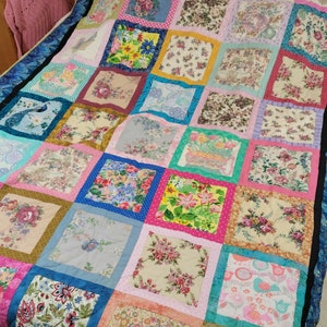 Könnte beinhalten: Eine Patchwork-Decke mit verschiedenen Blumenmustern in Rosa-, Blau-, Grün- und Gelbtönen. Die Decke besteht aus Quadraten mit unterschiedlichen Blumendesigns, die jeweils mit einem kontrastierenden Rand eingefasst sind.