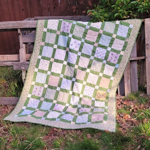 May include: A green and white patchwork quilt with a floral pattern. The quilt is made of squares of different fabrics, each with a different floral design. The quilt is folded and lying on a wooden fence.