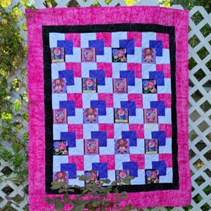 May include: A pink and white quilt with a black border. The quilt is made up of squares with a floral pattern. The quilt is hanging on a white lattice fence.