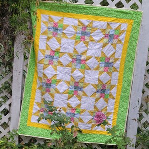Peut inclure: Une courtepointe blanche avec un motif de patchwork de étoiles jaunes, vertes et roses. La courtepointe est faite de carrés de tissu cousus ensemble pour créer un motif d'étoile. La courtepointe est accrochée à une clôture en treillis blanc.