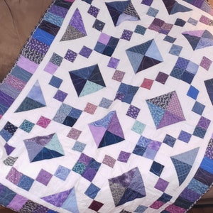 May include: A white quilt with a geometric pattern of purple, blue, and green squares and triangles. The quilt is edged with a purple and blue border.