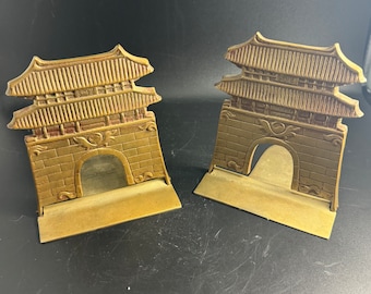 May include: A pair of gold-colored metal bookends shaped like traditional Asian pagodas. The bookends have a detailed design with intricate carvings and a layered roof.