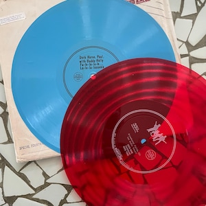 May include: Two vinyl records, one blue and one red, are stacked on top of a vintage record sleeve. The blue record has the text "Dark Horse, Paul, with Buddy Holly" and the red record has the text "Strawberry Fields #24".