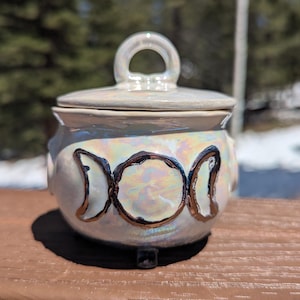 May include: Iridescent ceramic cauldron with a lid and handle. The cauldron features three crescent moon designs outlined in gold. The cauldron sits on three small feet. The background is blurred, with a brown surface in the foreground.