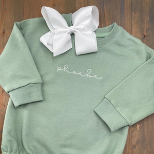 May include: A sage green baby onesie with long sleeves and a white bow on top. The onesie has the name "Phoebe" embroidered on the front.