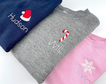 May include: Three folded sweatshirts in navy blue, gray, and pink. Each sweatshirt has embroidered details: a Santa hat with the name "Hudson", a candy cane with the name "Will", and a snowflake with the name "Ava".