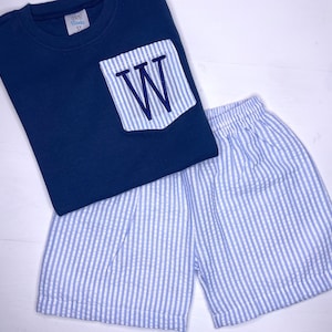 May include: A navy blue short-sleeved shirt with a white pocket featuring a blue embroidered letter "W". The shirt is paired with blue and white striped shorts.