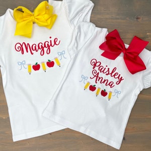 Op de afbeelding: Twee witte T-shirts met rode en gele borduursels van appels en potloden. De shirts zijn gepersonaliseerd met de namen "Maggie" en "Paisley Anna".