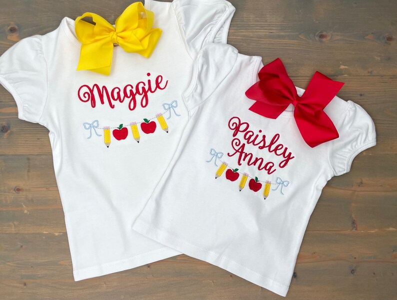Op de afbeelding: Twee witte T-shirts met rode en gele borduursels van appels en potloden. De shirts zijn gepersonaliseerd met de namen "Maggie" en "Paisley Anna".