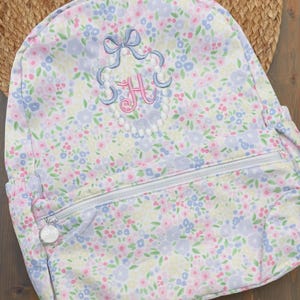 May include: A floral-patterned backpack in pastel colors, featuring a zipper closure and a decorative monogram with the letter "H". The backpack has a light blue bow and a pearl-like border around the monogram.