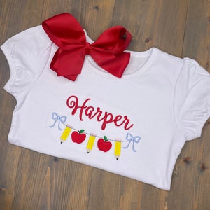 Op de afbeelding: Wit T-shirt met een rode strik erboven. Het shirt heeft een geborduurd ontwerp van een reeks potloden en appels met de naam "Harper" erboven.