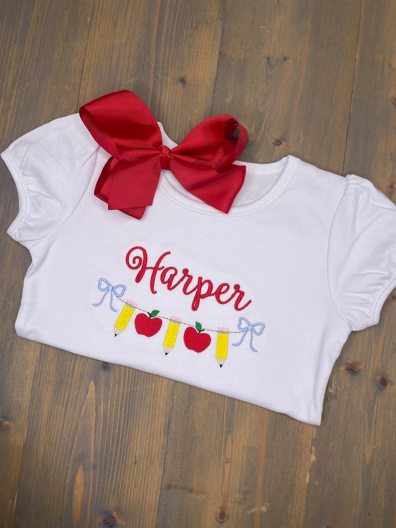 Op de afbeelding: Wit T-shirt met een rode strik erboven. Het shirt heeft een geborduurd ontwerp van een reeks potloden en appels met de naam "Harper" erboven.