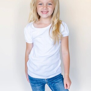 Op de afbeelding: Een jong meisje met blond haar draagt een wit shirt met korte mouwen en een blauwe jeans. Ze glimlacht en heeft een witte strik in haar haar.
