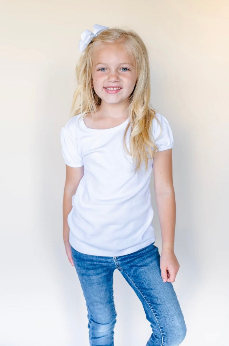 Op de afbeelding: Een jong meisje met blond haar draagt een wit shirt met korte mouwen en een blauwe jeans. Ze glimlacht en heeft een witte strik in haar haar.