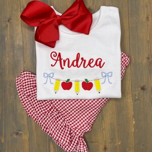 Op de afbeelding: Een wit T-shirt met de naam "Andrea" geborduurd erop. Het shirt heeft een rode strik bovenop en een rood-wit geruite rok eronder. Het shirt heeft ook een rij rode appels en gele potloden geborduurd erop.