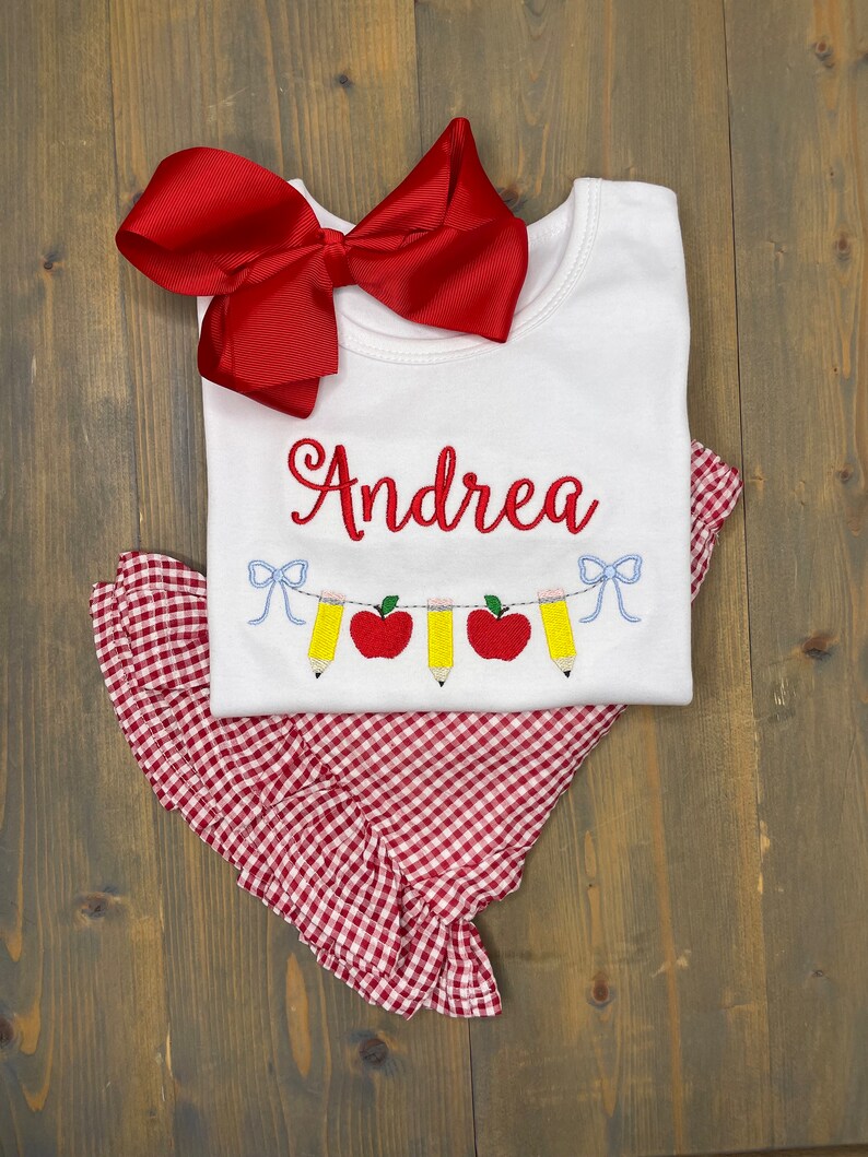 Op de afbeelding: Een wit T-shirt met de naam "Andrea" geborduurd erop. Het shirt heeft een rode strik bovenop en een rood-wit geruite rok eronder. Het shirt heeft ook een rij rode appels en gele potloden geborduurd erop.